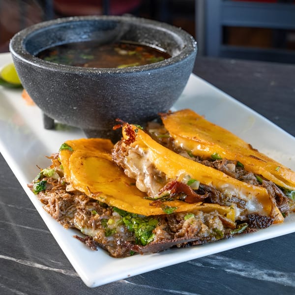 Birria tacos with melted cheese and cilantro, served with a side of consommé in a molcajete.