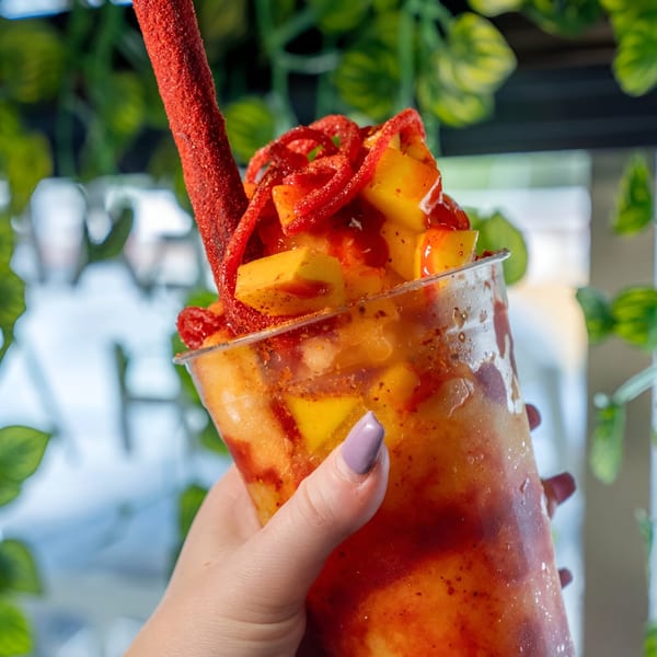 A hand holds a Mangonadas Mexican Kitchen drink with mango chunks, chamoy, and a tamarind straw.